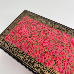Può includere: Una scatola rettangolare nera con un vivace motivo floreale rosa. I fiori sono raggruppati, creando un motivo denso. La scatola ha un bordo dorato con un design intricato e a spirale.