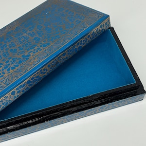 Peut inclure: Boîte rectangulaire ouverte, bleue à l'intérieur et à l'extérieur. Le couvercle est orné d'un motif floral doré. Les bords sont noirs et l'intérieur est bleu uni.