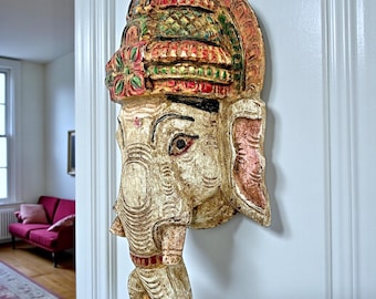 Peut inclure: Une tête d'éléphant en bois sculpté, accrochée au mur. La sculpture présente une coiffe détaillée avec des accents rouges, verts et dorés. Le visage de l'éléphant est peint dans des tons crème et rose, avec des sculptures complexes et une trompe incurvée.