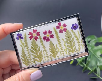 Op de afbeelding: Een rechthoekige, zilverkleurige etui met een witte binnenkant, met een bloemenontwerp. Het ontwerp bevat geperste bloemen in tinten roze, paars en groen, gerangschikt in een natuurlijk patroon. De etui wordt door een hand vastgehouden.