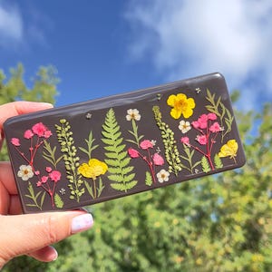 Pode incluir: Uma bandeja retangular cinza-escura com um arranjo decorativo de flores secas prensadas e folhas de samambaia verdes incrustadas na superfície. O design botânico inclui pequenas flores rosas, amarelas e brancas sobre a base escura.