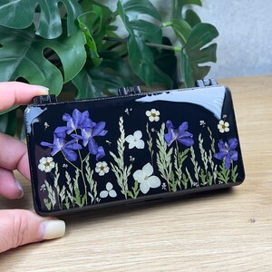 Pode incluir: Um estojo retangular preto com acabamento em resina brilhante incrustado com flores prensadas roxas e brancas, juntamente com folhagem verde. O design exibe um arranjo botânico de pequenas flores e caules finos sobre a superfície escura.
