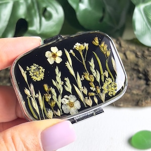 Pode incluir: Pequena caixa de comprimidos retangular com borda prateada e fundo preto. Decorada com flores silvestres e ervas amarelas e brancas secas, encapsuladas em resina transparente. A caixa é segurada por uma pessoa.