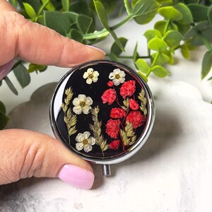 May include: A round, silver-toned pill box with a black background. The box is decorated with dried flowers in white, red, and yellow. The flowers are arranged in a decorative pattern. The pill box is held by a person.