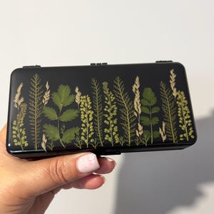 Pode incluir: Um estojo retangular preto com acabamento brilhante e uma disposição decorativa de folhas verdes prensadas e ervas secas na parte frontal. Os elementos botânicos estão dispostos num padrão vertical sobre o fundo escuro.