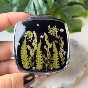 May include: A square, silver-framed compact mirror with a black background. The mirror's surface features a botanical design of pressed ferns, grasses, and small white flowers. The plants are arranged to create a natural, meadow-like scene. The compact is held in a hand.