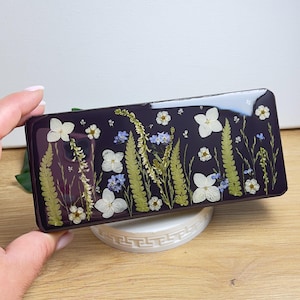 May include: A rectangular, dark purple resin case with a glossy finish. The case is adorned with pressed white, blue, and yellow flowers and green ferns. The case is resting on a white, decorative stand.