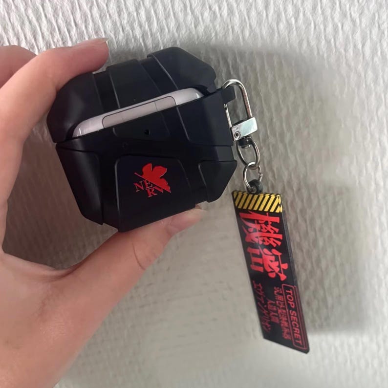 May include: Black protective case for wireless earbuds, with a red logo and a keychain tag with Japanese text and "TOP SECRET". The case is open, showing the white earbuds. The case is designed to protect the earbuds.