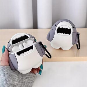 以下が含まれることがあります： グレーと白のサメ型AirPodケース。ケースは開いており、イヤホンが見えます。2つ目のケースが開いたケースの隣に表示されています。どちらにも、キーチェーンやバッグに取り付けるための黒いループが付いています。