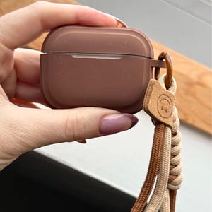 Może przedstawiać: Brązowe etui na AirPody z pasującym paskiem na nadgarstek. Etui ma jednolity, stonowany brązowy kolor. Pasek na nadgarstek wykonany jest z plecionych brązowych i beżowych sznurków, z małą skórzaną metką z uśmiechniętą buzią.