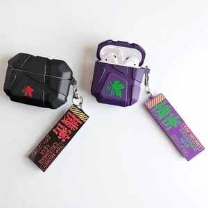 May include: Two AirPod cases, one black and one purple, with keychains. The black case has a red logo, the purple case a green logo and is open, showing white earbuds. The keychains have Japanese text and the words 'TOP SECRET'.