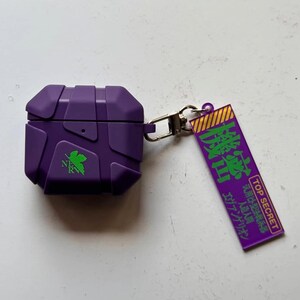 May include: A purple protective case for wireless earbuds, featuring a geometric design and green detailing. It includes a silver clasp and a purple tag with Japanese text and the words "TOP SECRET".