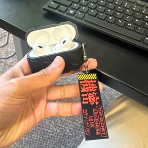 May include: Black protective case for wireless earbuds, open to reveal white earbuds. A black and yellow tag with Japanese text and "TOP SECRET" hangs from the case. The text on the tag includes "Evangelion" and "Artificial Human".