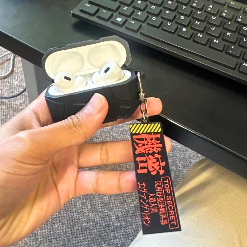 May include: Black protective case for wireless earbuds, open to reveal white earbuds. A black and yellow tag with Japanese text and "TOP SECRET" hangs from the case. The text on the tag includes "Evangelion" and "Artificial Human".