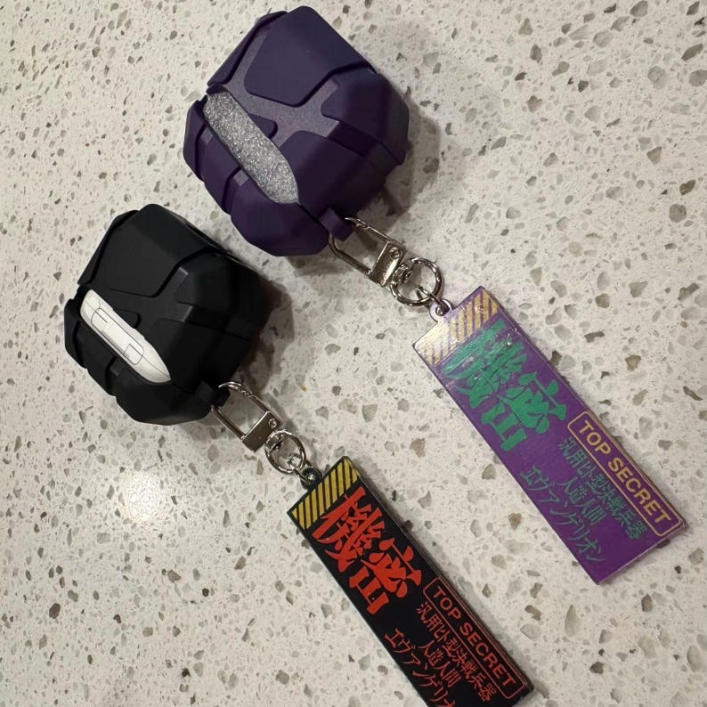 May include: Two protective cases for wireless earbuds, one black and one purple, with silver keychains. Each case has a rectangular tag with Japanese text and the words "TOP SECRET". The purple case has a grey interior.