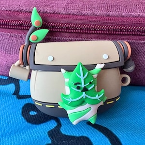 以下が含まれることがあります： 緑の葉の形をしたキャラクターと植物のデザインが施された、ベージュとグレーのケース。ケースは円筒形で、ハンドルと葉の付いた植物の茎が付いています。緑のキャラクターは白と緑のストライプのデザインです。