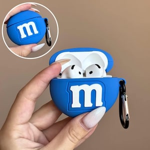 Może przedstawiać: Niebieskie silikonowe etui na AirPods z białym logo "m". Etui jest otwarte, odsłaniając białe słuchawki AirPods. Mniejszy, okrągły obraz zamkniętego etui znajduje się w lewym górnym rogu. Dołączony jest czarny karabińczyk.