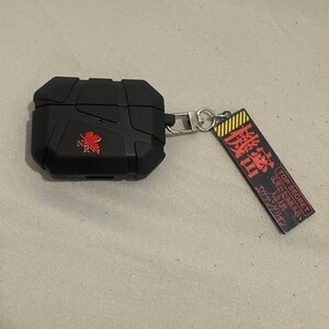 May include: Black protective case for wireless earbuds with a red and white logo. The case has a rugged design and includes a keychain with Japanese text and the words "TOP SECRET".