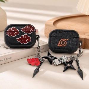 May include: Two black protective cases for wireless earbuds. One case features red cloud patterns, while the other displays an orange leaf symbol and embossed text. Both cases include metal keychains with charms shaped like clouds, stars, and blades.