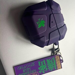 May include: A purple earbud case with a geometric design and a green logo. It has a matching purple tag with Japanese text and a silver-coloured clasp. The case is designed to protect wireless earbuds.