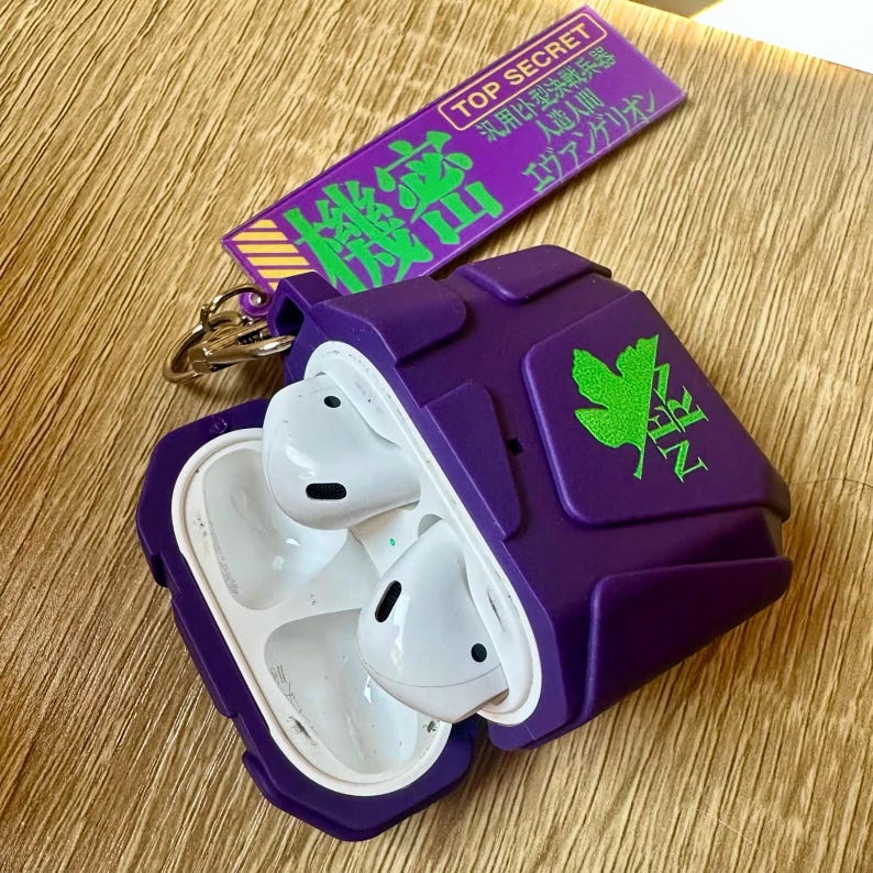 May include: A purple protective case for wireless earbuds, featuring a green logo and Japanese text. The case is open, showing white earbuds. A matching purple and green keychain tag with Japanese text is also visible.