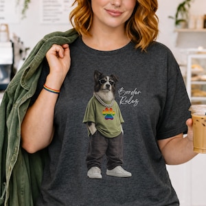 Puede incluir: Camiseta gris oscuro con un perro con gafas de sol, una camisa verde con una huella de pata arcoíris y pantalones grises. El texto "Border Rules" está escrito en la camisa. La persona de la imagen lleva una chaqueta verde y sostiene una taza de café.