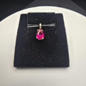 May include: A gold pendant with a vibrant, teardrop-shaped, pink gemstone. The pendant is displayed on a black velvet surface, set against a white background. The gemstone is the focal point, showcasing its color and shape.