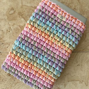 Könnte beinhalten: Eine rechteckige Laptophülle aus gehäkeltem, dickem Stoffgarn in Pastellfarben wie Rosa, Hellblau, Gelb und Lavendel. Die Hülle ist mit einem kleinen beigen Stoffetikett versehen, auf dem der Text "tinku crochet" aufgedruckt ist.