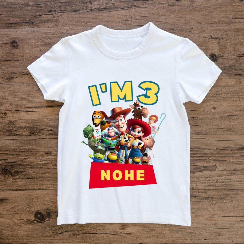 May include: White t-shirt featuring the phrase "I'M 3" in yellow and blue, with a red banner that says "NOHE". The shirt has a colorful graphic of characters from the Toy Story movie.