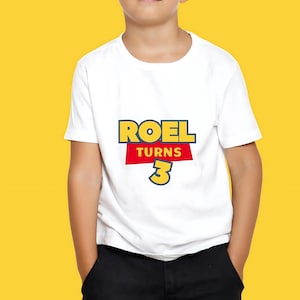 May include: White t-shirt with the text "ROEL TURNS 3" in a colorful, playful design. The text is in a bold, cartoon-like font, with a yellow and red color scheme. The shirt is worn by a child.