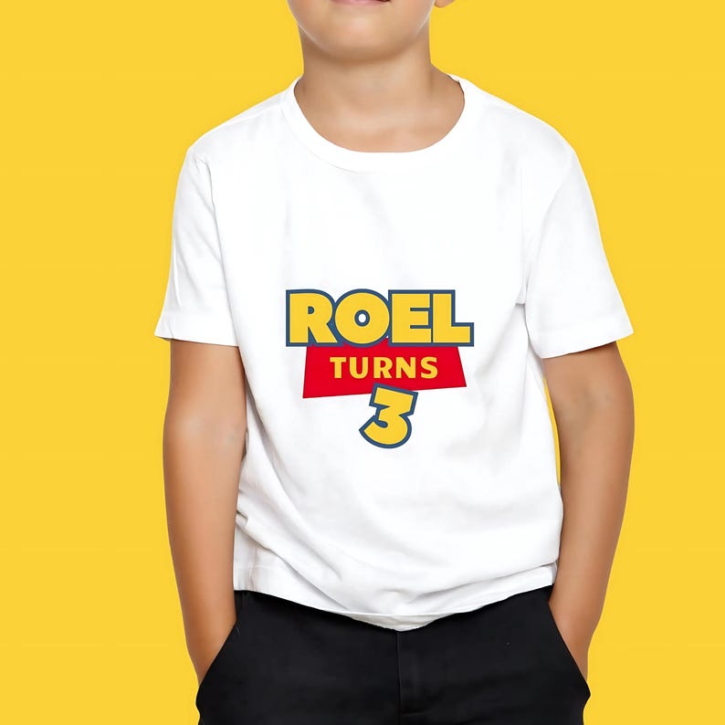 May include: White t-shirt with the text "ROEL TURNS 3" in a colorful, playful design. The text is in a bold, cartoon-like font, with a yellow and red color scheme. The shirt is worn by a child.