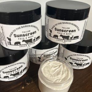 May include: Four jars of homemade tallow sunscreen with a white label that reads "Back to the Basics Homestead Tallow Sunscreen Face & Body Cocoa Butter 4 oz". One jar is open and shows the white, creamy texture of the sunscreen.