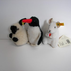 May include: Three plush animal toys: a cream-colored pug with black spots, a black and white penguin with a red beak, and a white rabbit holding a red heart. Each toy has a tag.