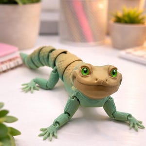 May include: A detailed, articulated toy lizard with a tan head and segmented back, and light green limbs. The lizard has bright green eyes and is posed on a white surface. The toy is a fun, decorative item.