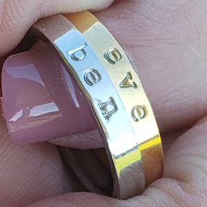 May include: A gold and silver ring with the word "be love" engraved on the band. The ring is held up against a green background.