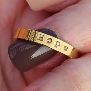 May include: A gold-toned metal ring featuring a smooth, polished band with the word Hope stamped in black, capital and lowercase letters across the front surface. The ring has a simple, flat design with a consistent width throughout the band.