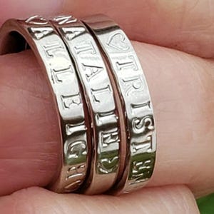 May include: Three stacked silver rings with a polished finish. Each band features engraved capital letters and heart symbols. The visible text on the rings includes the names TRISTEN, NATALIE, and ALLEIGH, with heart icons placed between the names.