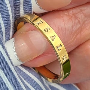 May include: A gold-toned metal ring with a flat, polished surface featuring the engraved text PSALM. The band has a smooth, uniform width and a reflective finish, showcasing clear, capital letters stamped into the metal along the outer circumference.