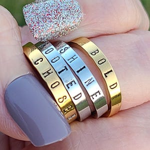 May include: Stack of four rings in gold and silver tones, each engraved with a word. The words are "CHOSE", "ROOTED", "SHINE", and "BOLD". The rings are on a hand with polished nails.
