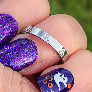 May include: A silver-toned metal ring featuring a repeating engraved pattern of small witch hats around the outer band. The ring has a smooth, polished finish and a flat profile, showcasing a consistent design of multiple hats encircling the band.