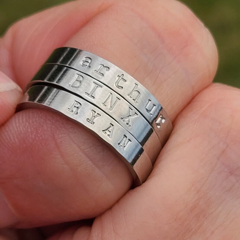 May include: Three silver-colored, stackable rings with engraved names: "arthur", "BINX", and "RYAN". The rings are worn on a finger, showcasing the personalized inscriptions. The rings are made of a shiny, metallic material.