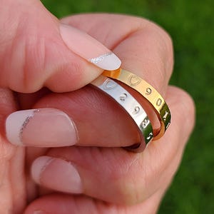 May include: Three thin, engraved rings in gold and silver tones. The rings feature numbers and a heart-shaped design. The rings are held between fingers with a green background.