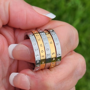 May include: A set of four stacked metal rings in alternating silver and gold tones. Each ring features engraved lowercase lettering. The visible text on the rings includes the words bibliophile, bookworm, bookish, and book nerd, arranged in a vertical stack.