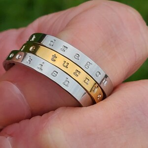 May include: A stack of three metal rings featuring stamped lowercase lettering. The top and bottom rings are silver-toned, while the middle ring is gold-toned. The visible text on the rings includes the letters d, r, a, g, o, n, t, u, r, n, i, s, h.