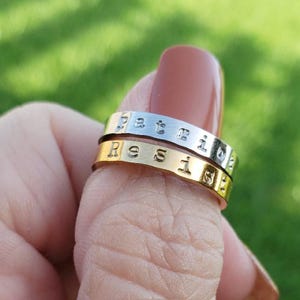 May include: Two personalized rings, one silver and one gold, stacked on a finger. The silver ring has the word "Patricia" stamped on it, and the gold ring has the word "Ressie" stamped on it. The background is a blurred green.