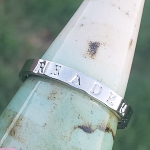 May include: A silver-toned metal ring featuring the stamped letters R, A, D, and B along its outer band. The ring is positioned on a tapered, light-colored, textured object. The surface of the ring is smooth and reflective with clearly visible indentations.