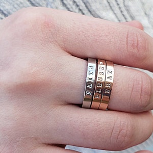 May include: Three stackable rings in silver, rose gold, and gold tones. Each ring has a stamped word: "FAITH", "BLESSED", and "PRAY". The rings are displayed on a person's finger.