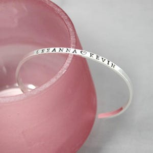 May include: A silver-colored bracelet with the names "LEEANNA" and "KEVIN" stamped on it, separated by a heart symbol. The bracelet is curved and rests against a pink, translucent background.