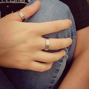 May include: A close-up of a person's hand wearing two silver rings. The rings are simple bands with engraved lettering. The hand is resting on a pair of blue jeans.