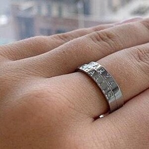 May include: A silver-colored metal ring featuring two stacked bands. The top band is engraved with the name ARYA and the bottom band is engraved with the name CHARLIE. The ring has a smooth, polished finish and a simple, clean design.